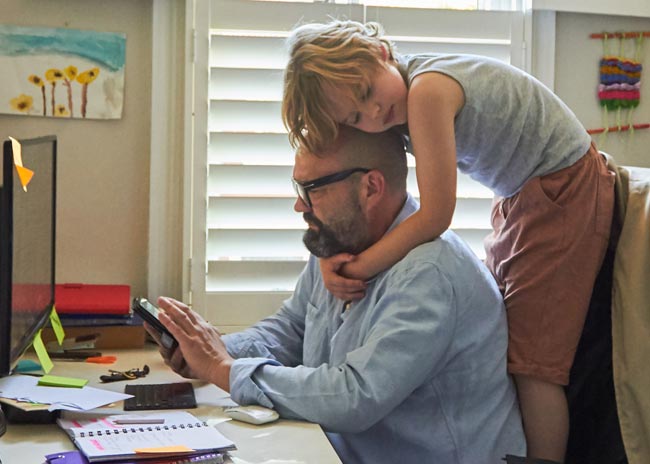 Man looking at mobile with child cuddling his neck.